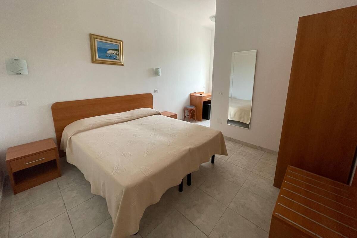 A bedroom with a bed, a nightstand, a desk, a chair, a mirror, and a framed picture on the wall.