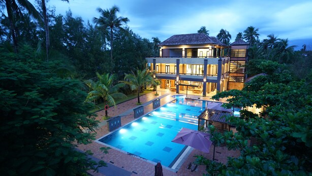 Um edifício de dois andares com piscina, cercado por vegetação exuberante e palmeiras.
