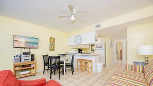 A living room with a red sofa, a TV, and a ceiling fan. There is a kitchen area with a clock and a dining table with chairs. - *WEEKLY SPECIALS!* * Island Winds 1BR! (Gulf Shores)