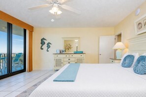 A bedroom with a large bed, a ceiling fan, a dresser, a mirror, and a view of the outside.