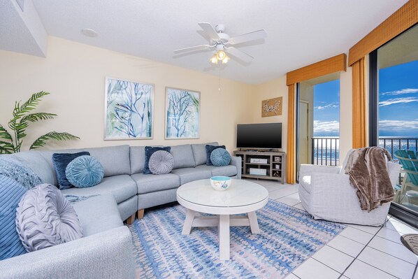 A living room with a grey sofa, a round coffee table, and a flat-screen TV. There is a large window with a view of the ocean.