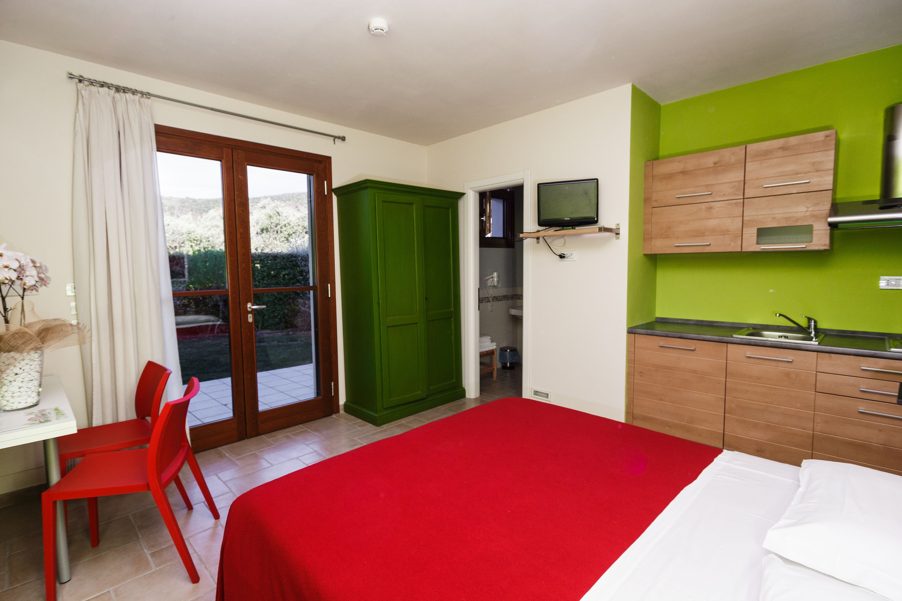 A bedroom with a red bedspread, a green wardrobe, a small kitchen with a sink, and a TV mounted above the counter.