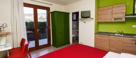 Een slaapkamer met een rode sprei, een groene garderobe, een kleine keuken met een spoelbak en een televisie die boven het aanrecht is bevestigd.