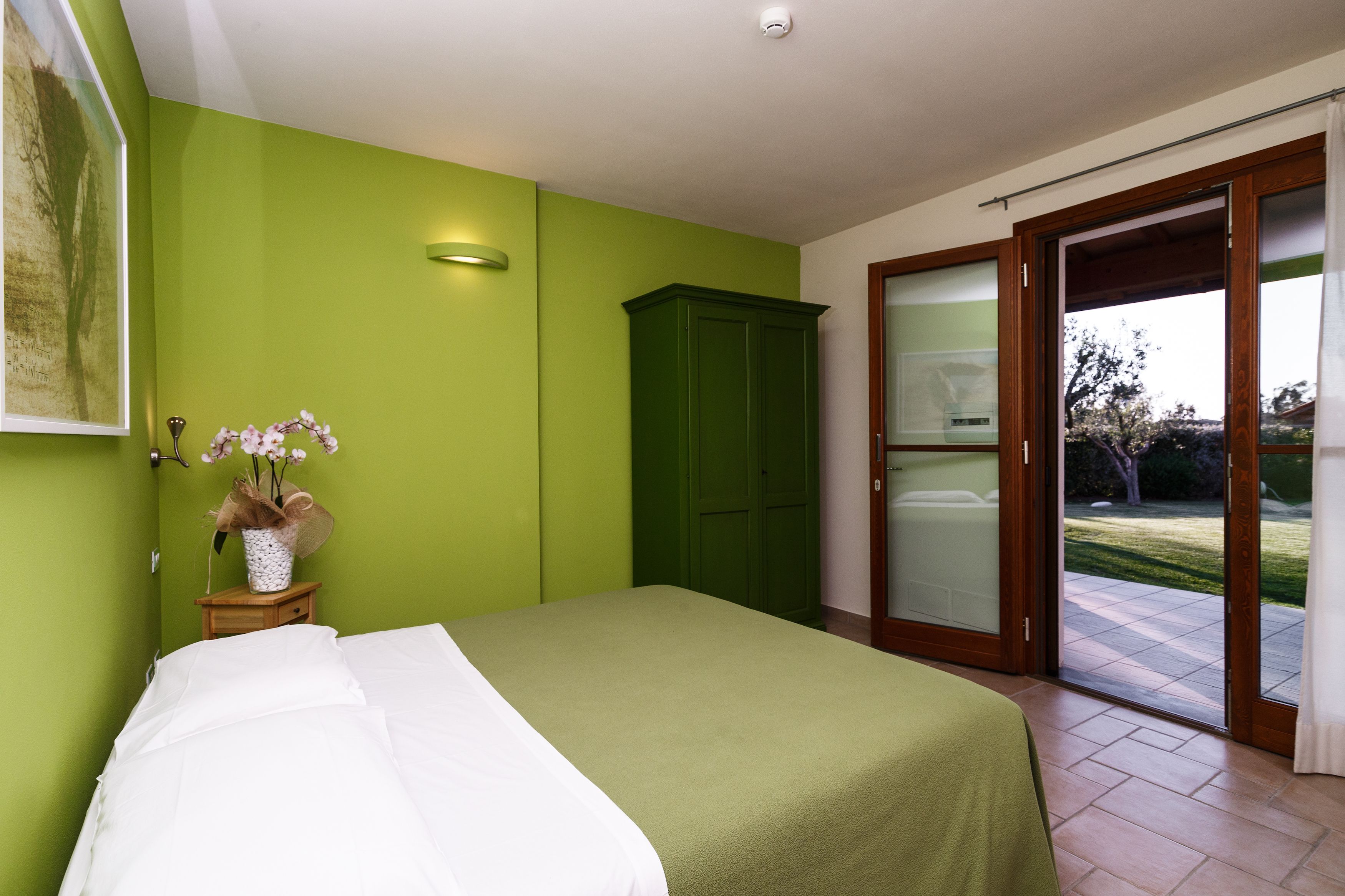 A bedroom with a bed, a green wall, a wooden cabinet, and a sliding glass door leading to an outdoor area.