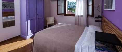 A bedroom with a bed, a purple wardrobe, a chair, a window with curtains, and a wall-mounted shelf.
