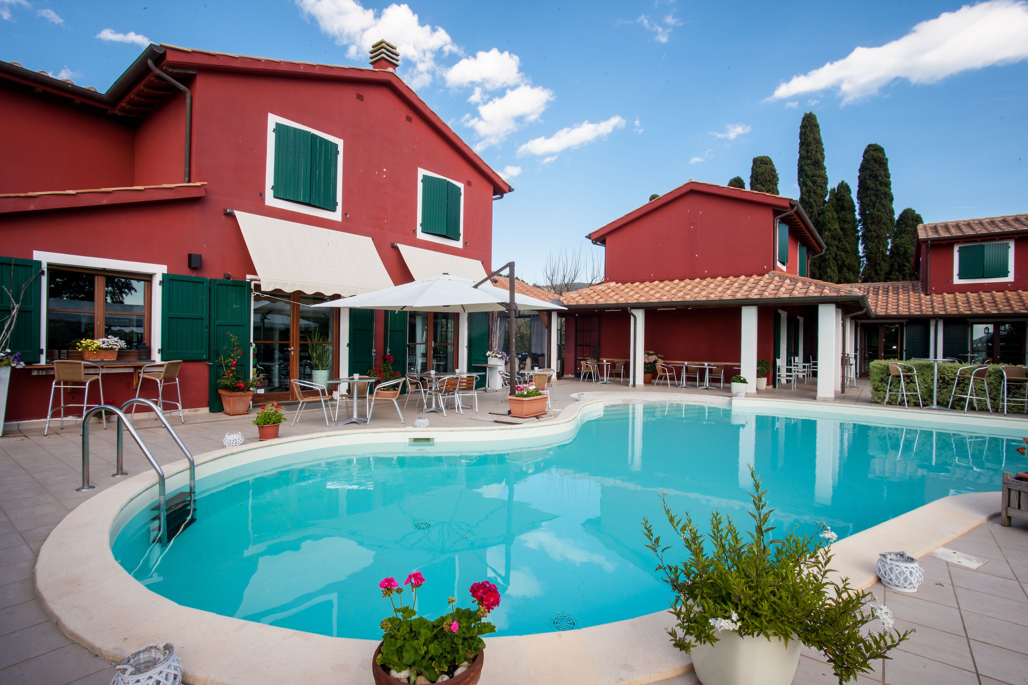 A swimming pool surrounded by red buildings with green shutters, outdoor seating, and potted plants.