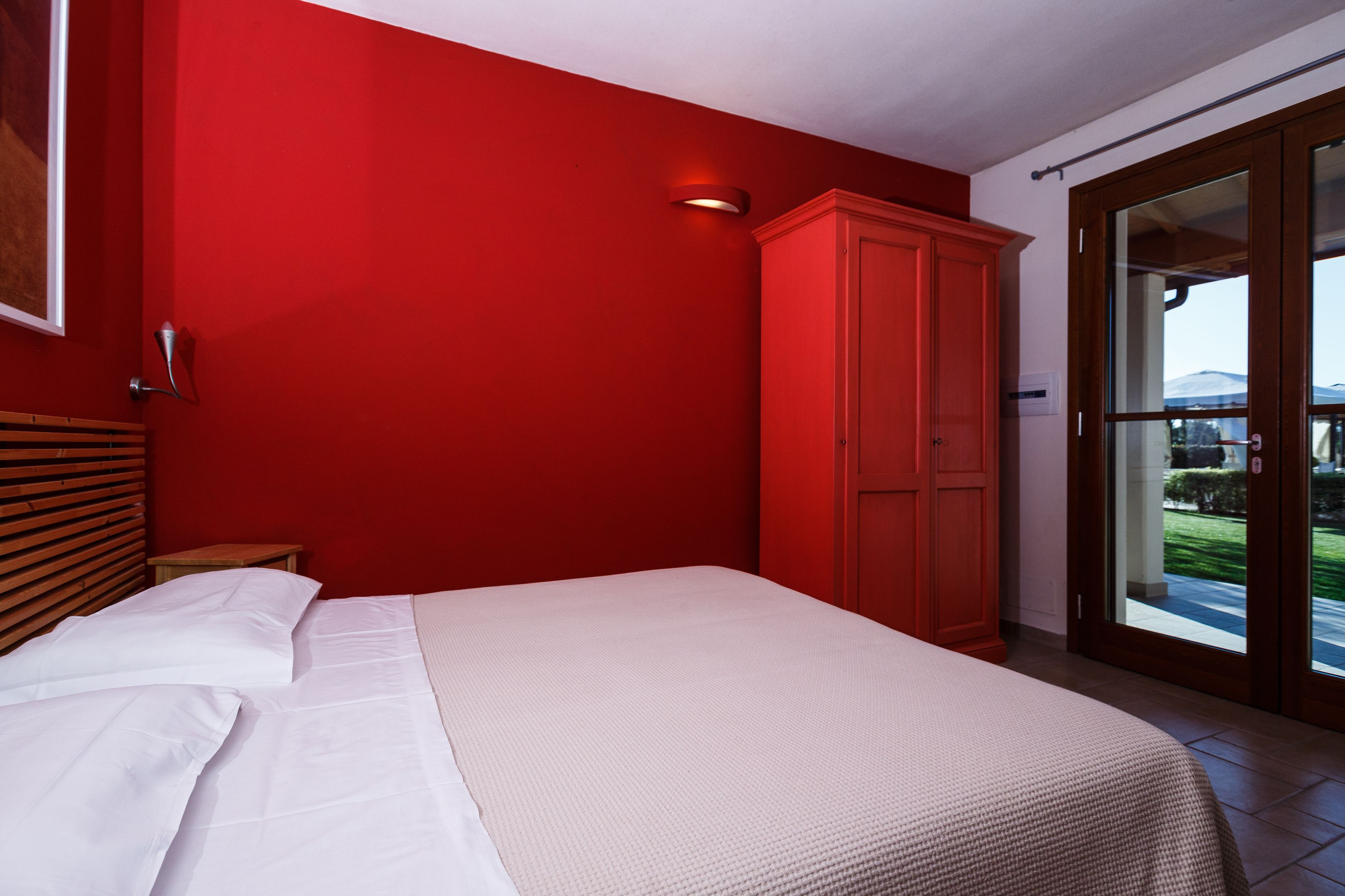 A bedroom with a bed, a red wall, and a sliding glass door leading to a balcony.