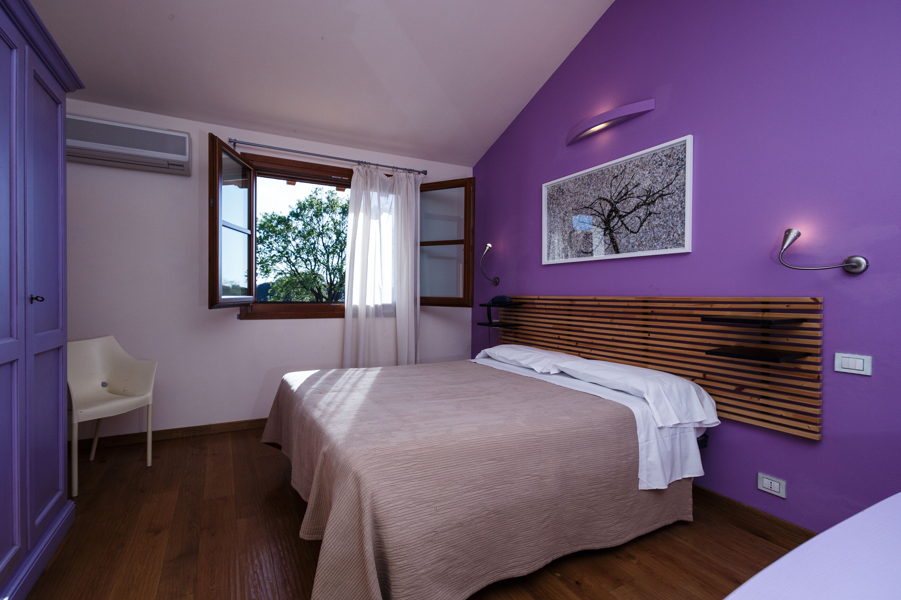 A bedroom with a bed, a chair, a window with curtains, a wall-mounted light, and a picture.