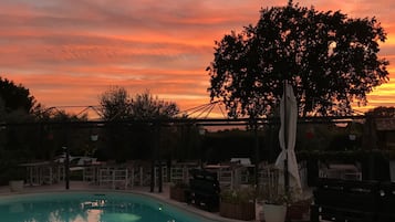 A swimming pool with a sunset view, surrounded by outdoor furniture and a large tree.