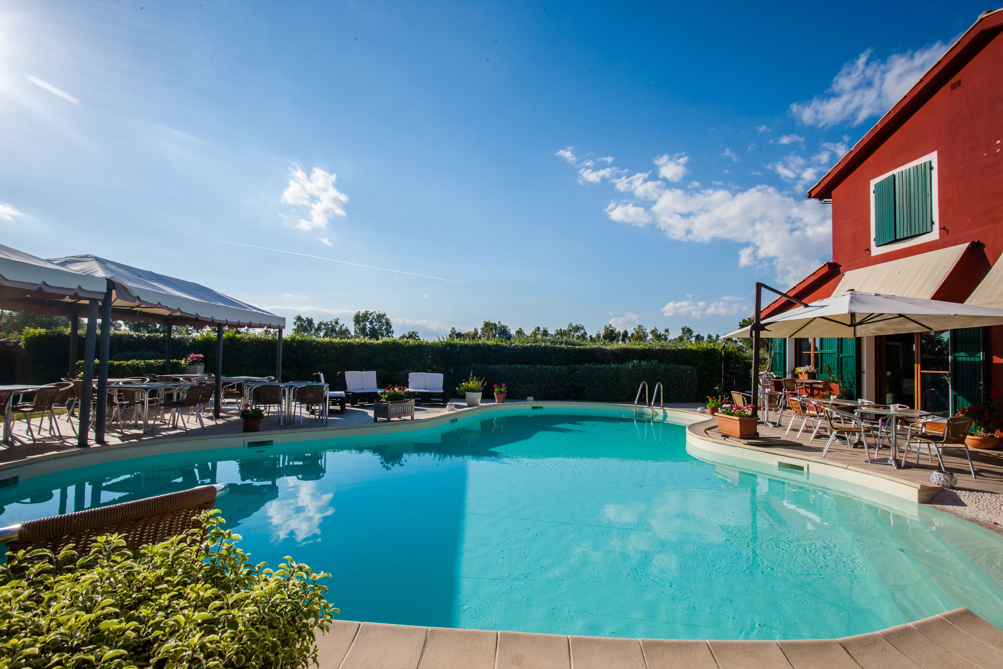 A swimming pool with lounge chairs and umbrellas, surrounded by a red building and a covered area with tables and chairs.