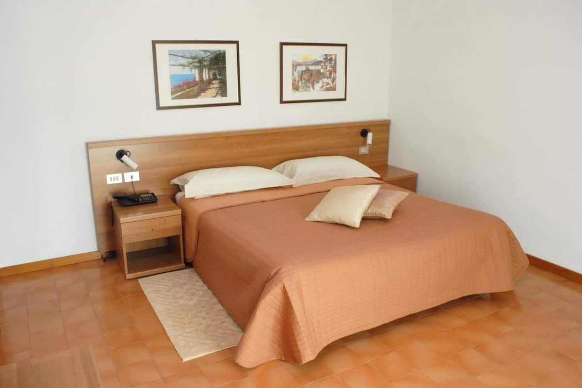 A bedroom with a wooden headboard, a bed with a peach-colored bedspread, two pillows, a bedside table with a lamp, and a nightstand. There are two framed pictures on the wall.