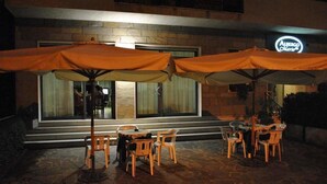 Un área de estar al aire libre con sombrillas naranjas y sillas de madera frente a un edificio con un gran ventanal y un letrero de neón.
