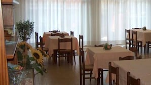 Un comedor con varias mesas y sillas, dispuesto en una sala amplia con ventanas grandes y cortinas.
