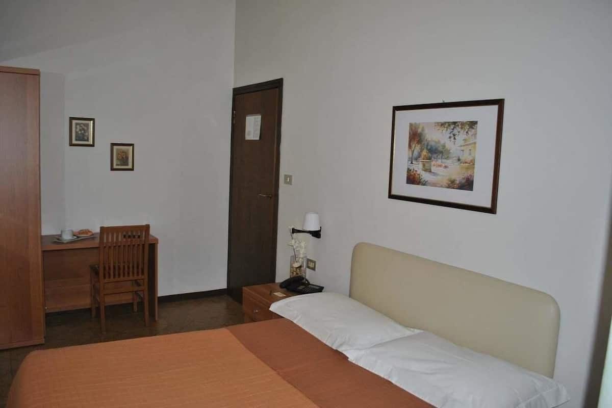 A hotel room with a bed, a desk with a chair, a nightstand with a lamp, and a door.