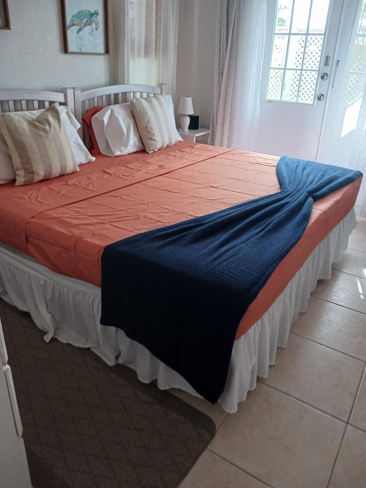 Un lit bien fait, avec une couvre-lit à motifs et un plaid bleu, une lampe de chevet et une porte donnant sur une fenêtre.