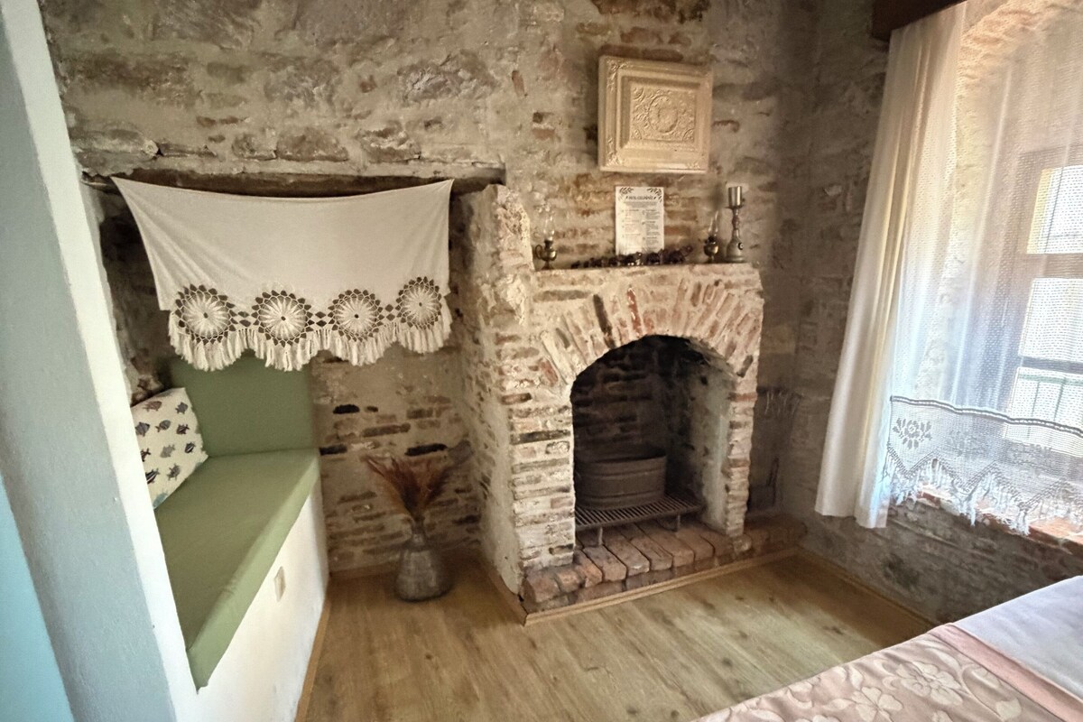 Une pièce rustique avec un mur en pierre, une cheminée, un banc recouvert d’une housse en dentelle et un lit agrémenté d’une couvre-lit à motifs floraux.