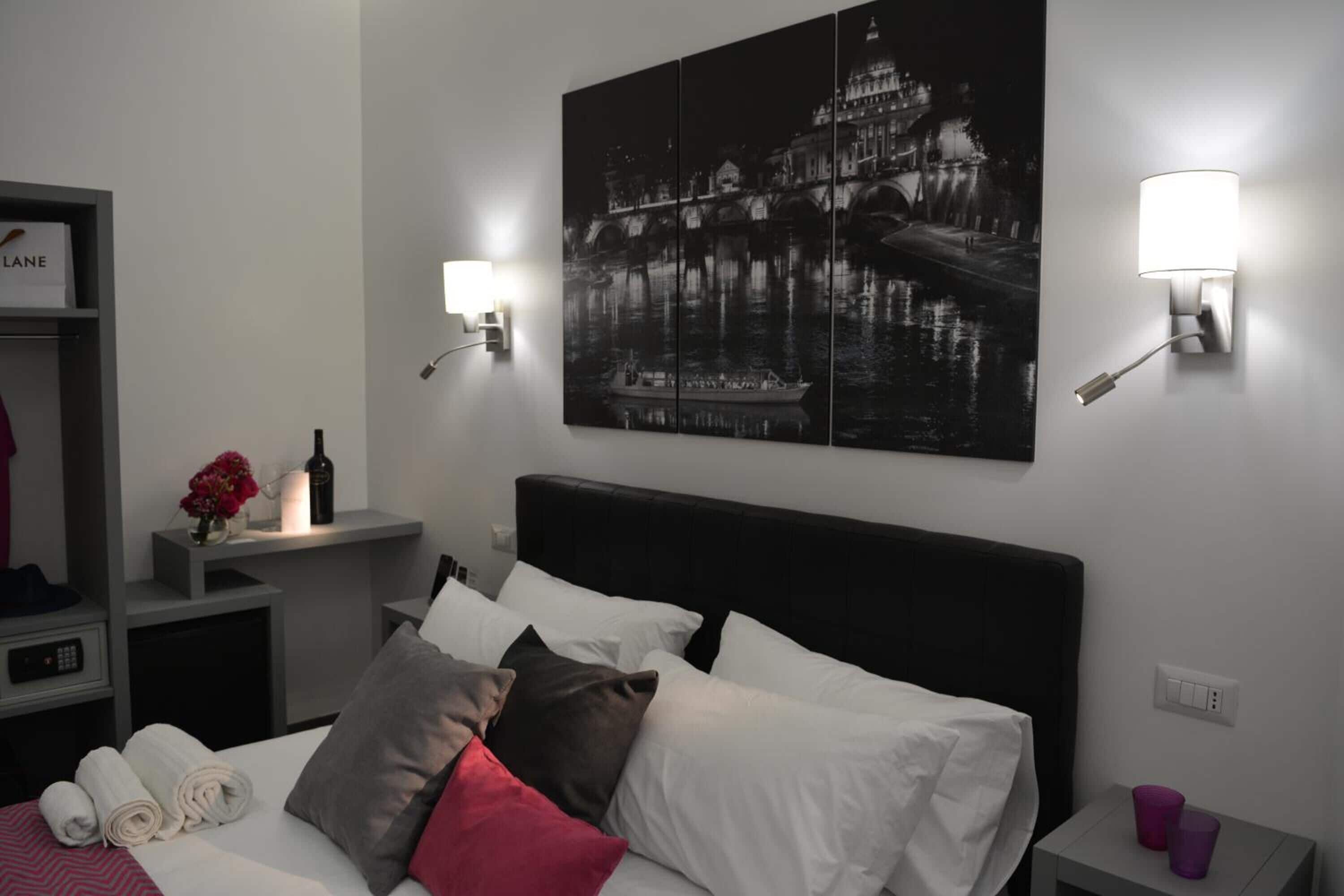 A hotel room with a bed, a nightstand, a wall-mounted lamp, and a framed picture of a cityscape.
