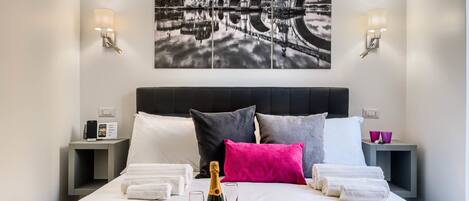 A hotel room with a bed, pillows, a bottle of champagne, glasses, and a tray of cherries.