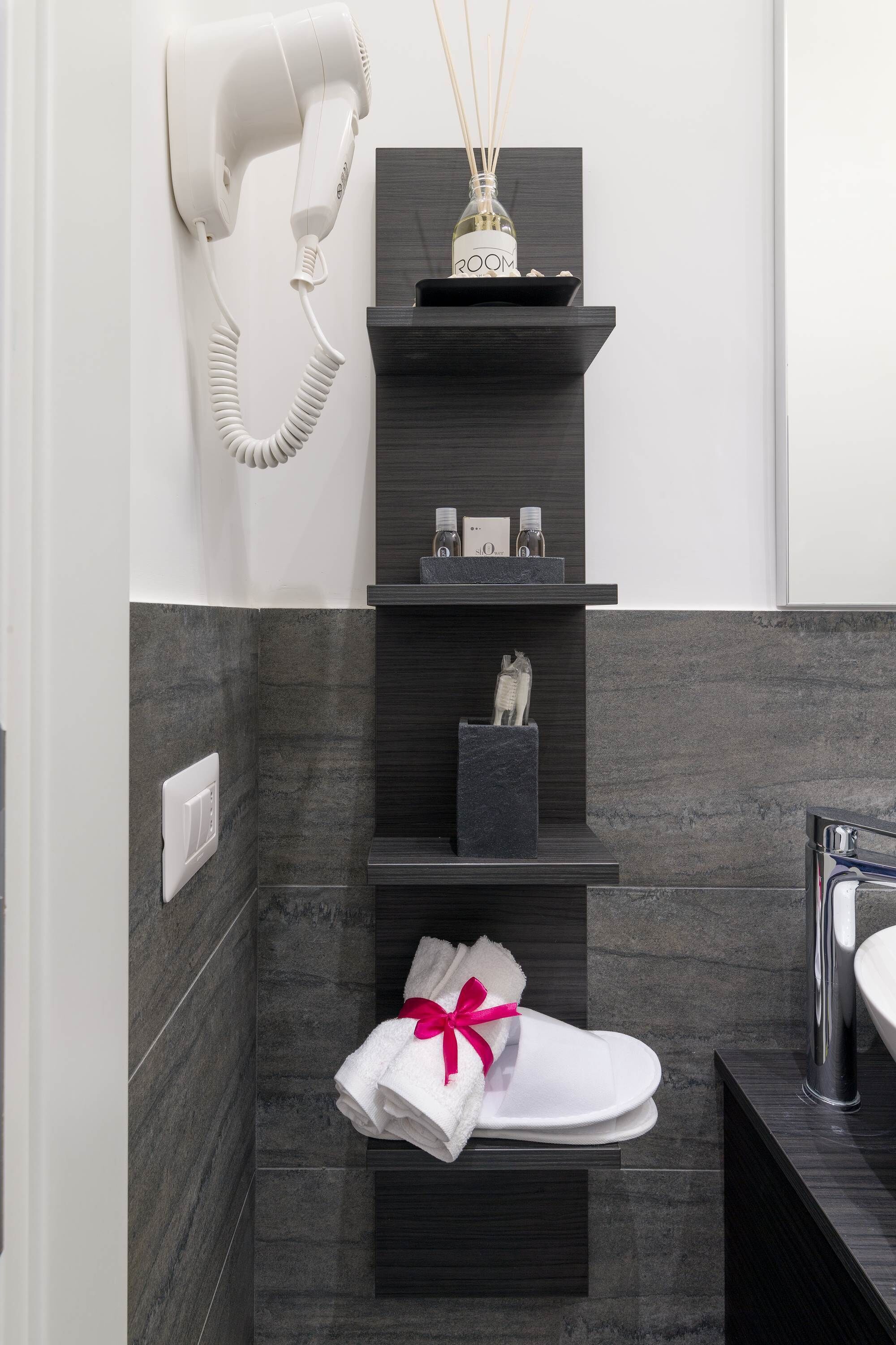 A modern bathroom with a hair dryer mounted on the wall, a shelf with toiletries, a towel rack with a towel and pink ribbon, and a dark marble wall.