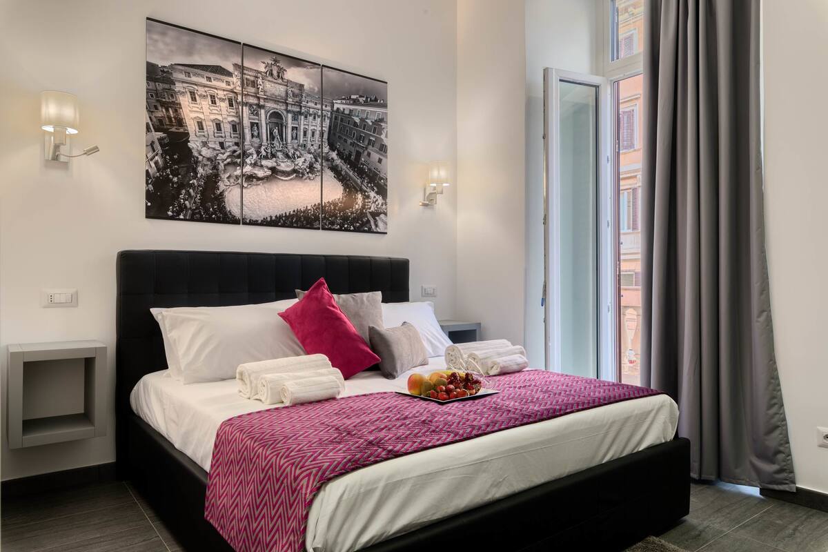 A neatly made bed with white linens and a pink patterned throw, a fruit bowl on the bed, and a cityscape painting above it.
