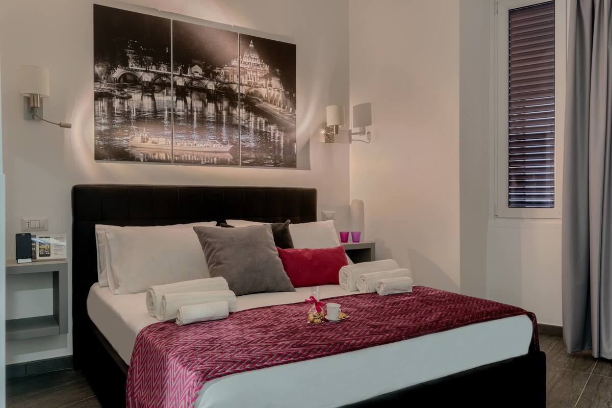 A hotel room with a large bed, a night scene artwork, bedside lamps, a nightstand, and a window with blinds.