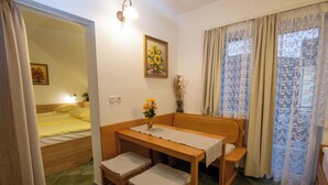 Una habitación pequeña con cama, una mesa de madera con dos sillas, un banco y una ventana con cortinas estampadas.
