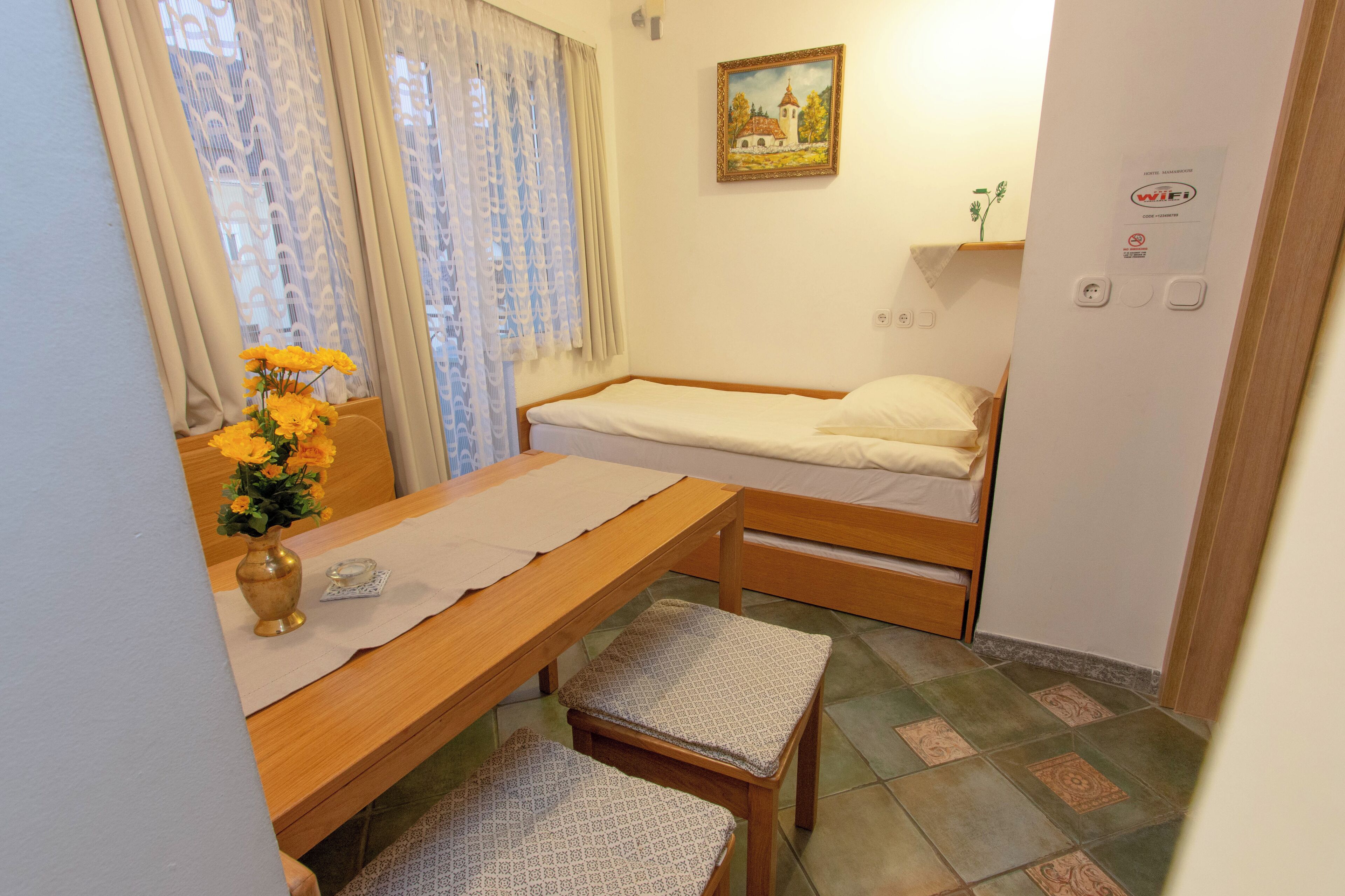 Quarto pequeno e arrumado com uma cama de solteiro, uma mesa de madeira com um vaso de flores, uma cadeira e um quadro na parede.