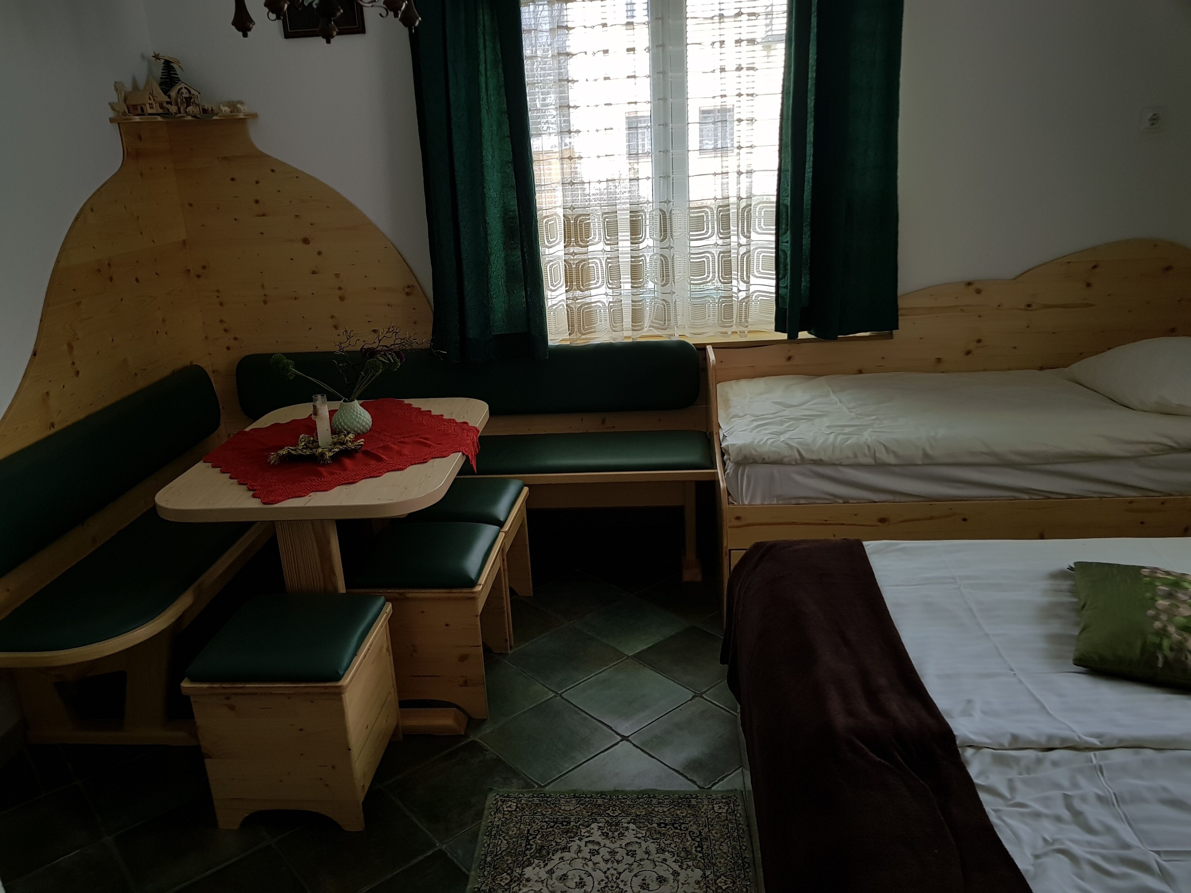 A cozy room with a wooden bed, a small table with a red cloth, a bench, and a window with green curtains.