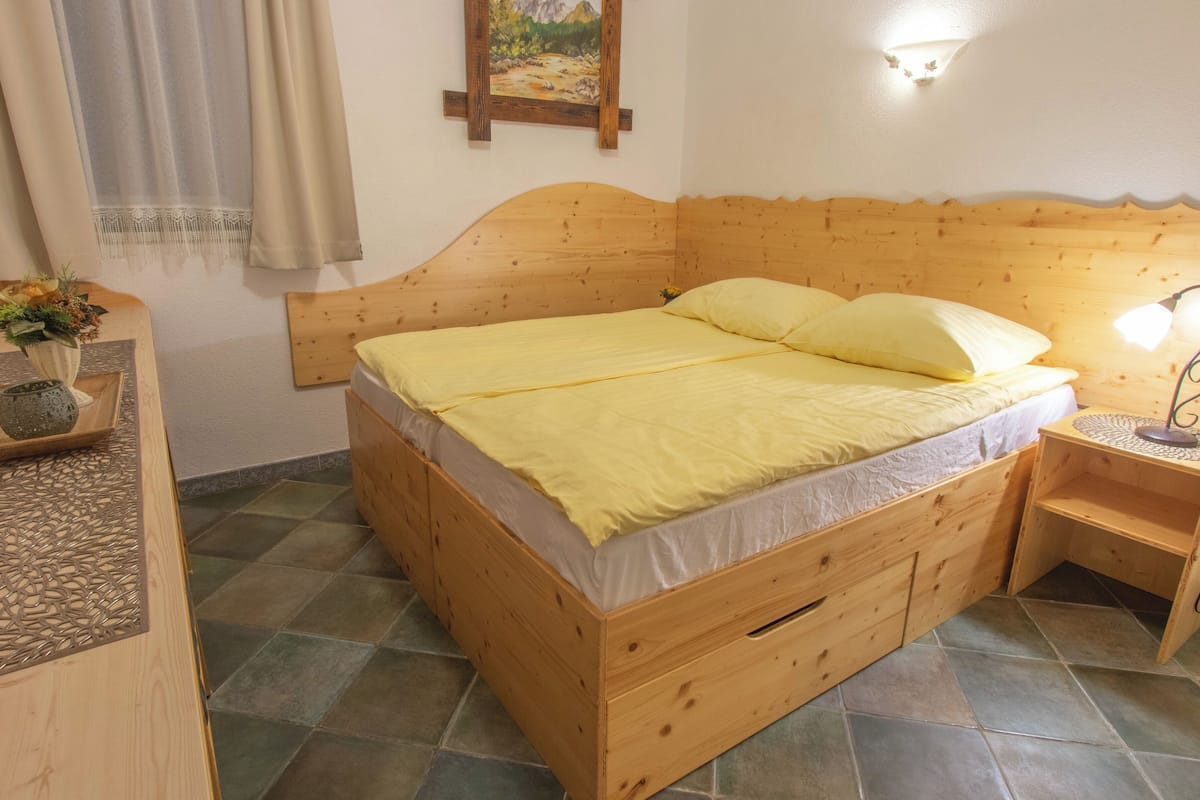 A wooden bed with a yellow bedspread and pillows, a bedside table with a lamp, and a painting on the wall.