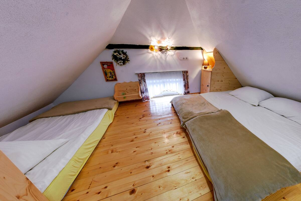 A cozy attic bedroom with two beds, wooden flooring, a small window, and a wall-mounted light fixture.