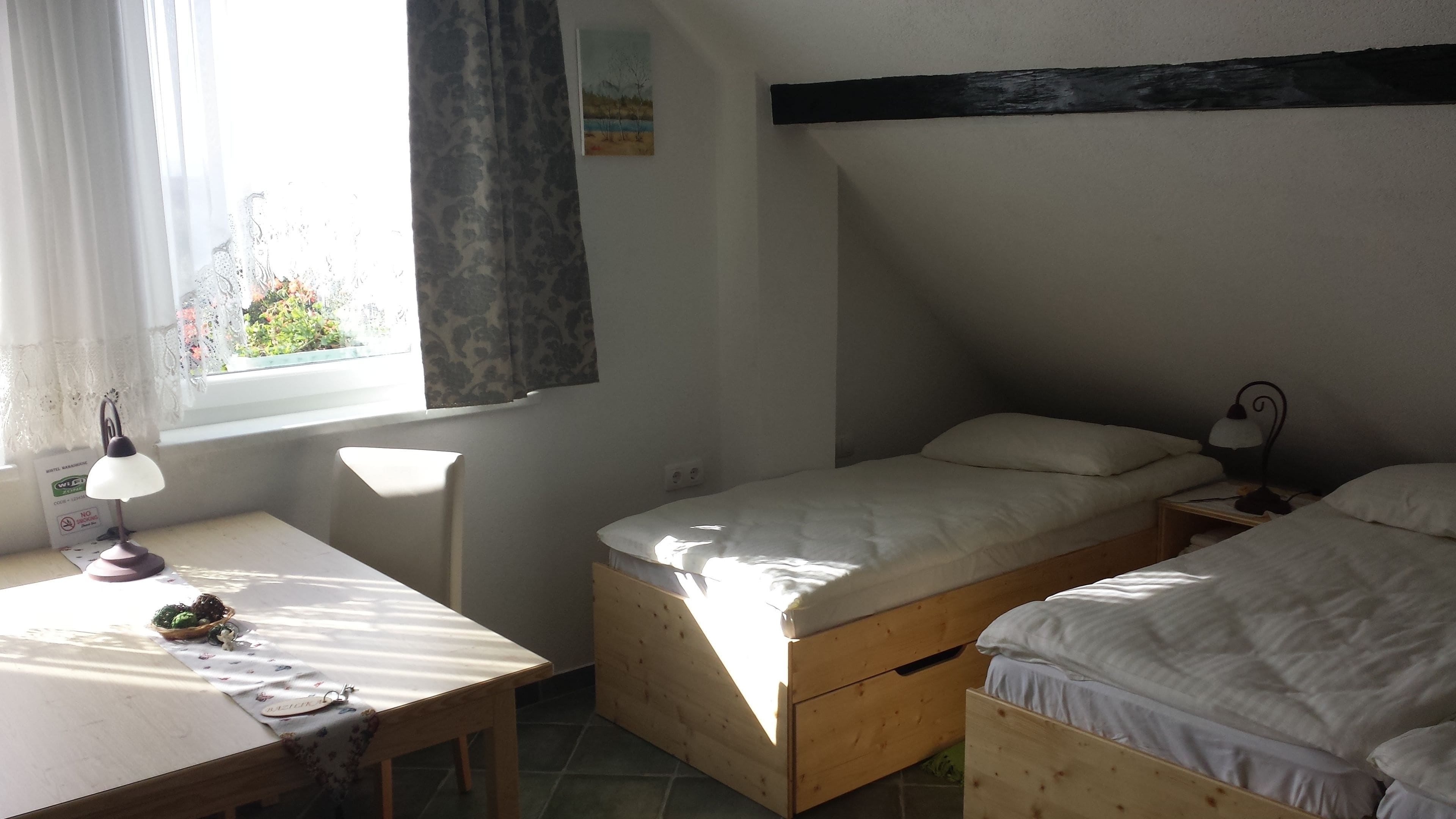 A small, cozy room with a desk, two single beds, a window with curtains, and a wall-mounted lamp.