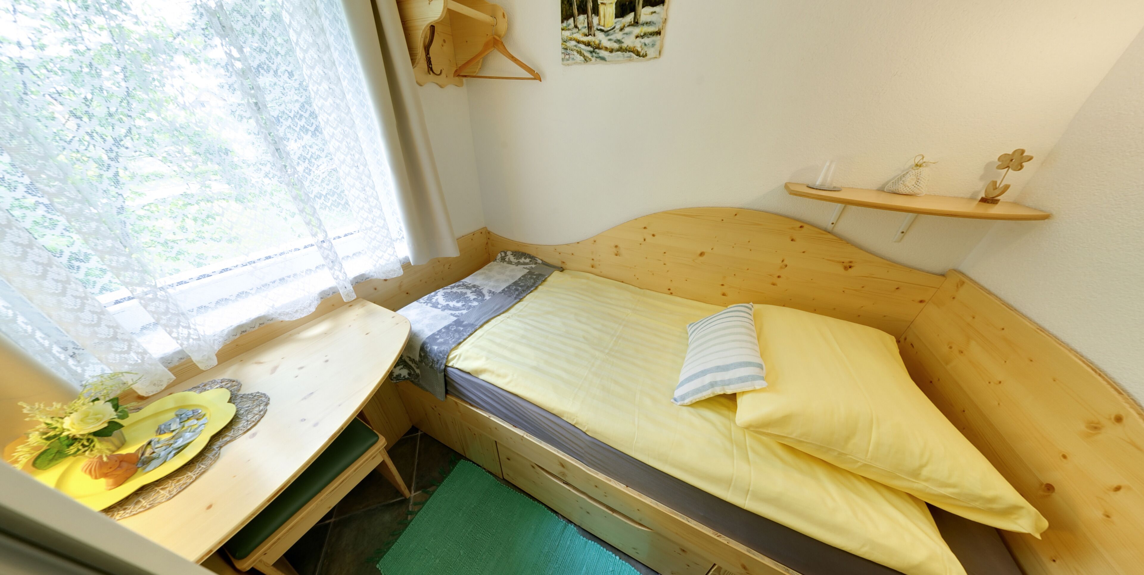 Une petite pièce agréable et chaleureuse, avec un lit simple, un bureau en bois et une fenêtre ornée de rideaux en dentelle.