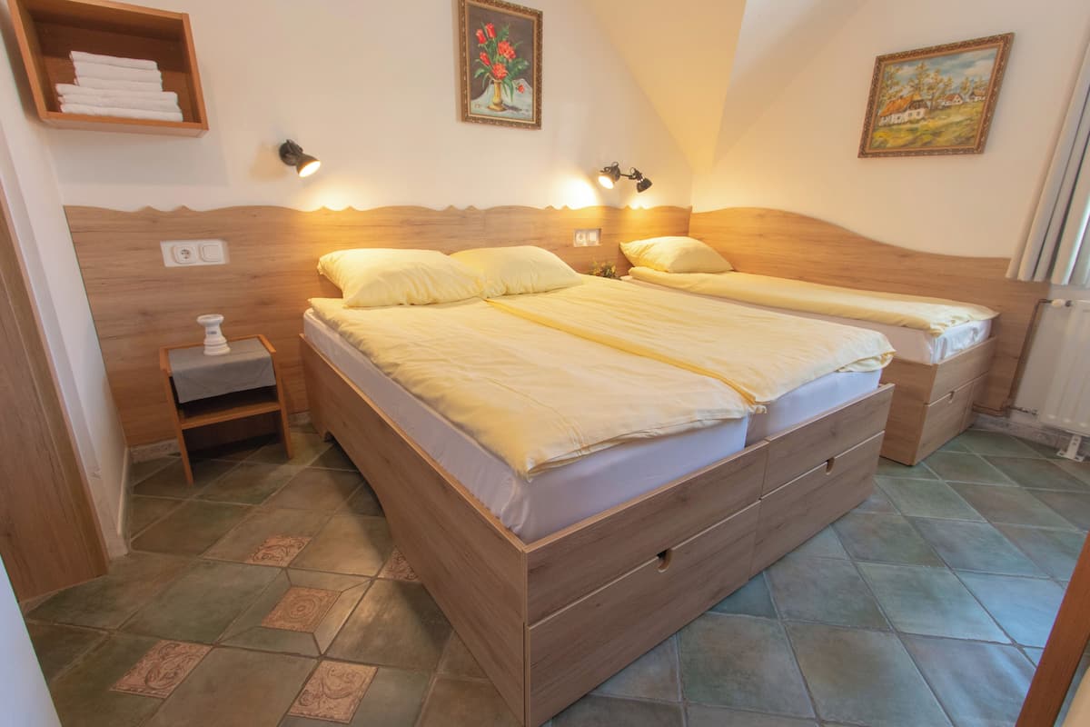 A bedroom with a wooden headboard, two beds with matching bedding, a nightstand with a lamp, a small shelf with towels, and a painting on the wall.
