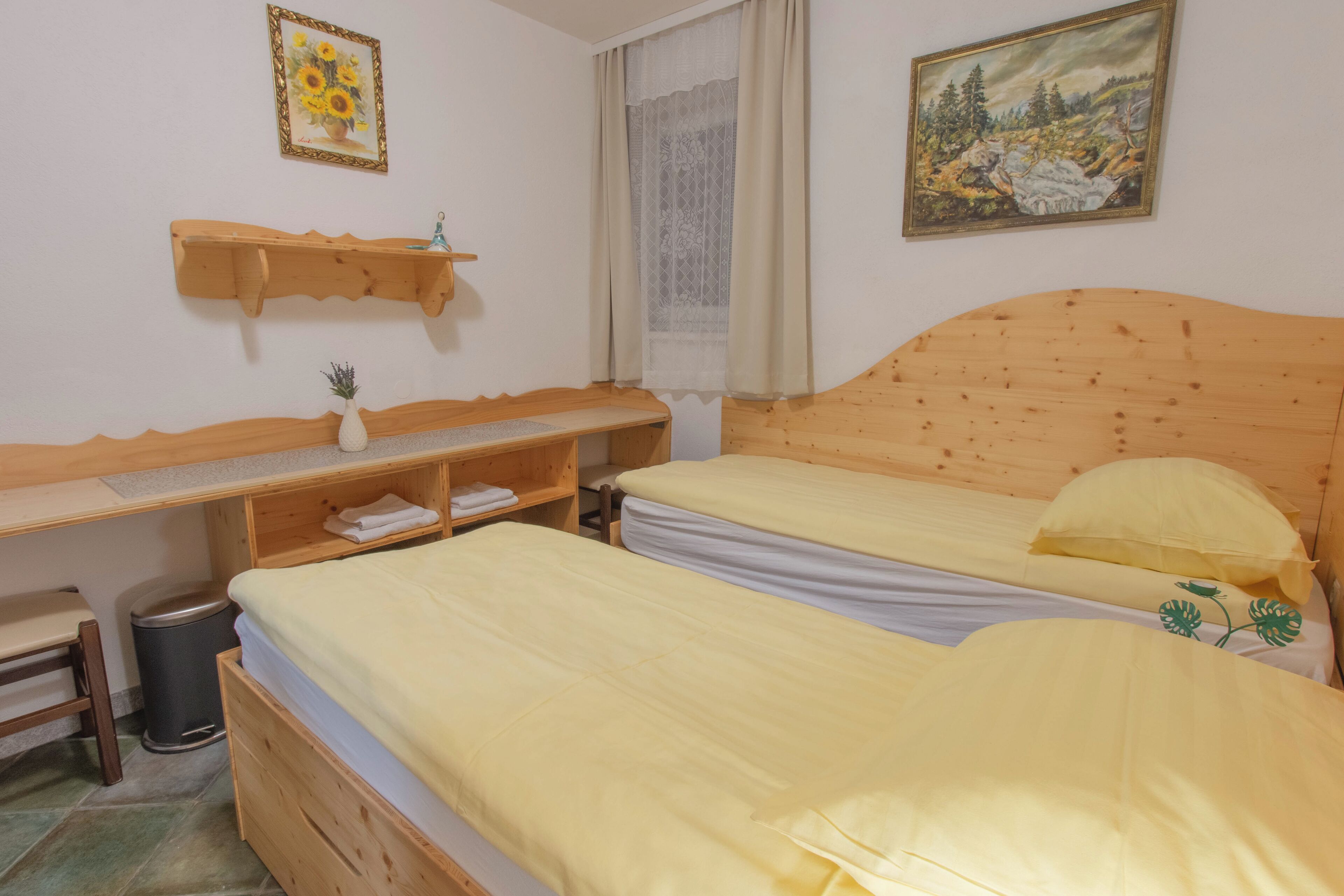 Una habitación con dos camas de madera, un escritorio de madera con silla, un estante pequeño y un cubo de basura.