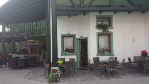 Ein Außenbereich mit Tischen und Stühlen, eine überdachte Terrasse und ein Gebäude mit grün gerahmten Fenstern.