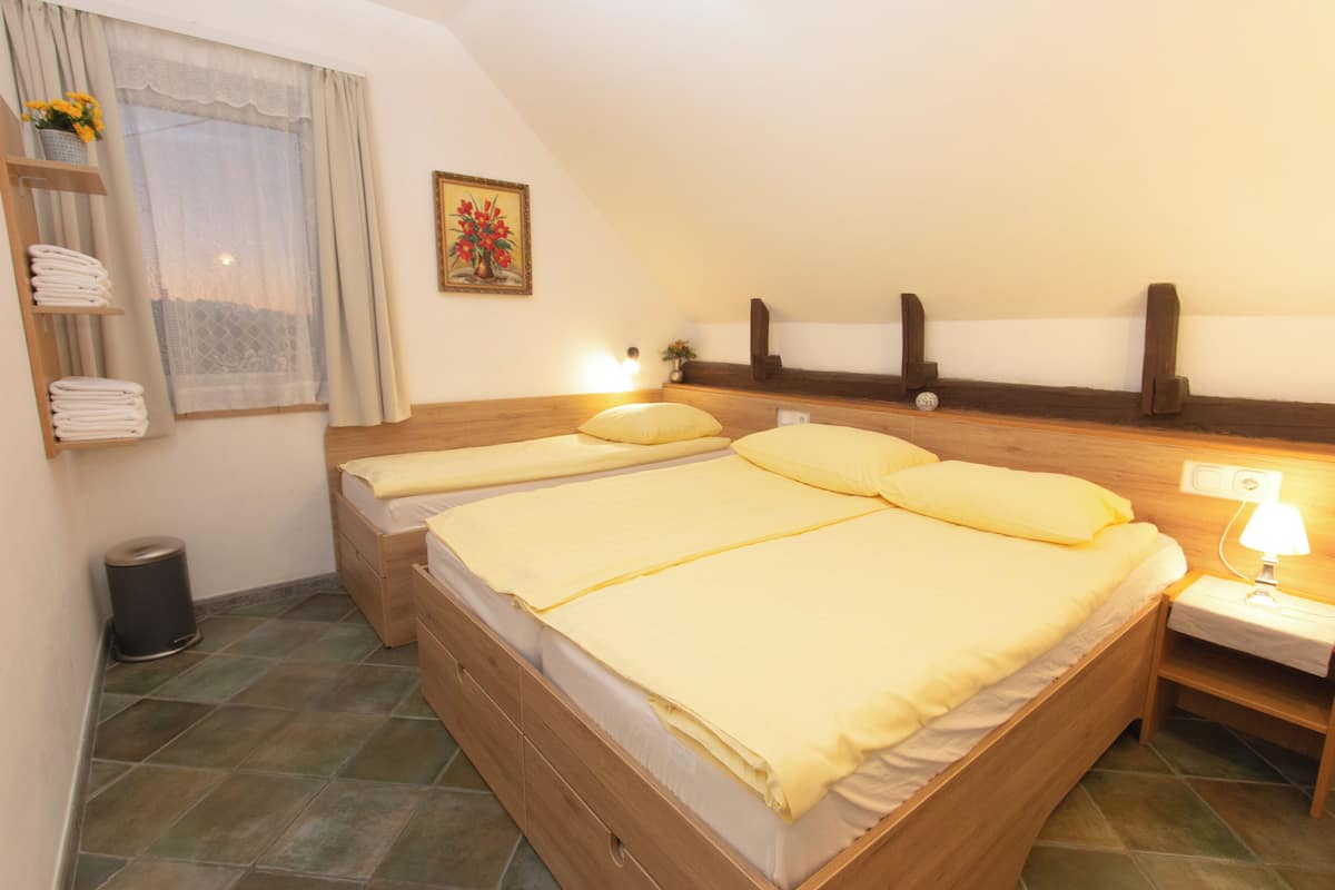 A bedroom with two beds, a window with curtains, a painting, a nightstand with a lamp, and a shelf with towels.
