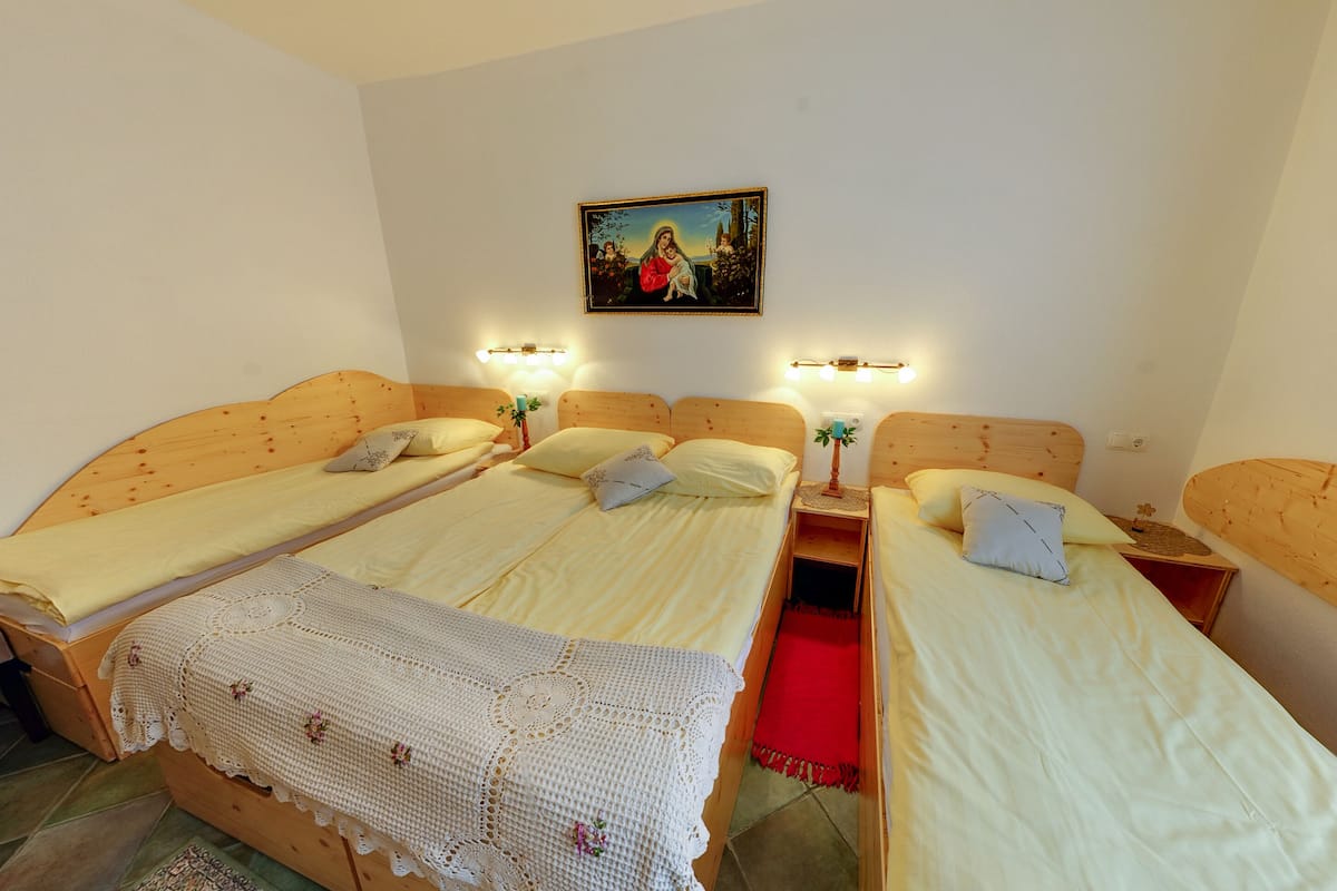 A room with three wooden beds, each with a pillow and a bedside table. There is a wall painting above the beds and a smaller painting on the wall to the right.