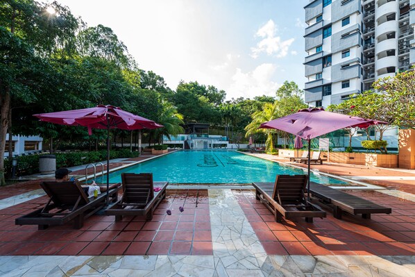 A swimming pool with lounge chairs and umbrellas, surrounded by greenery and a multi-story building. - Spacious Three Bedroom Condo Cyberjaya (Cyberjaya)