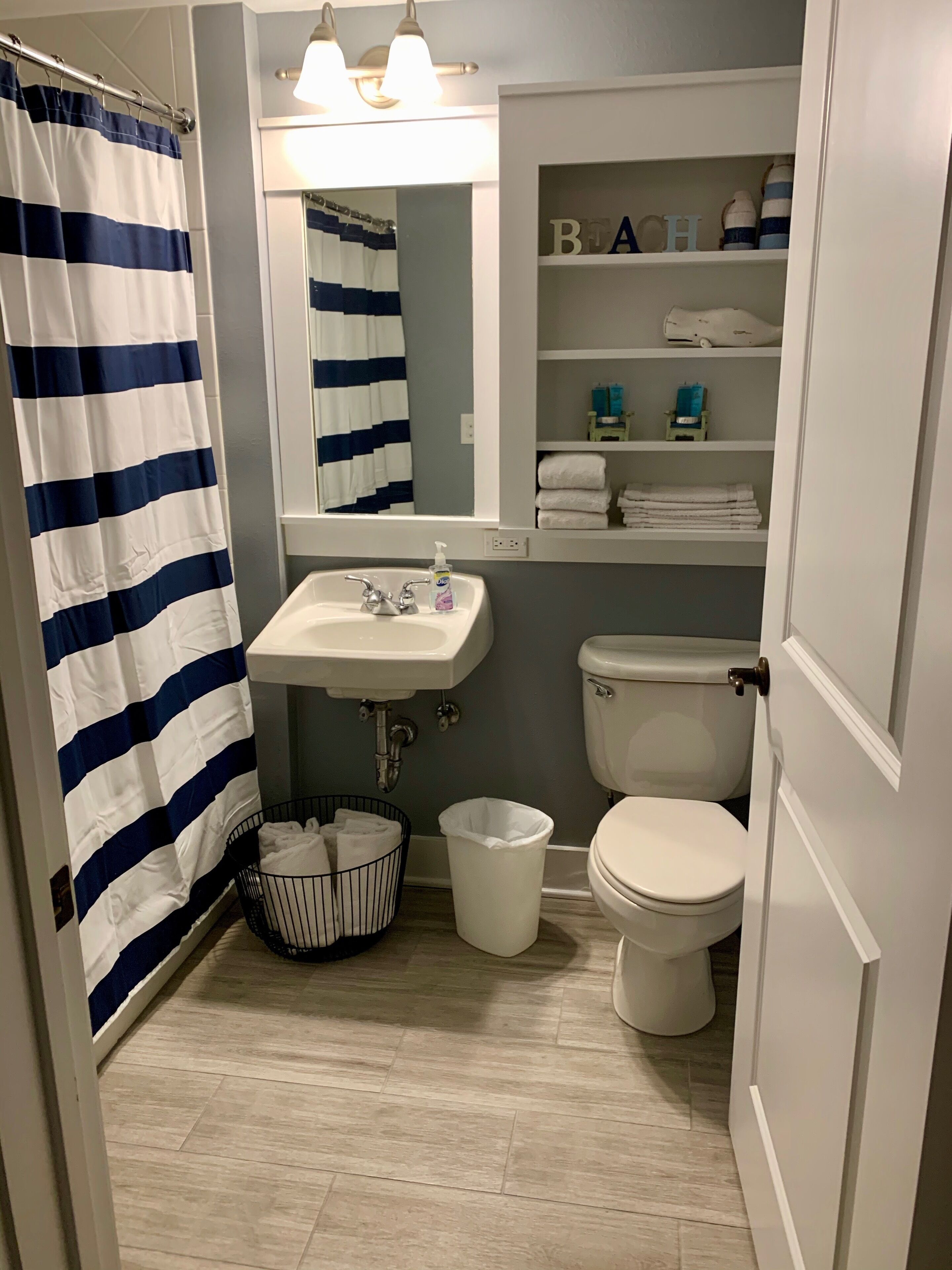 A bathroom with a striped shower curtain, a sink, a toilet, a mirror, and shelves with toiletries.