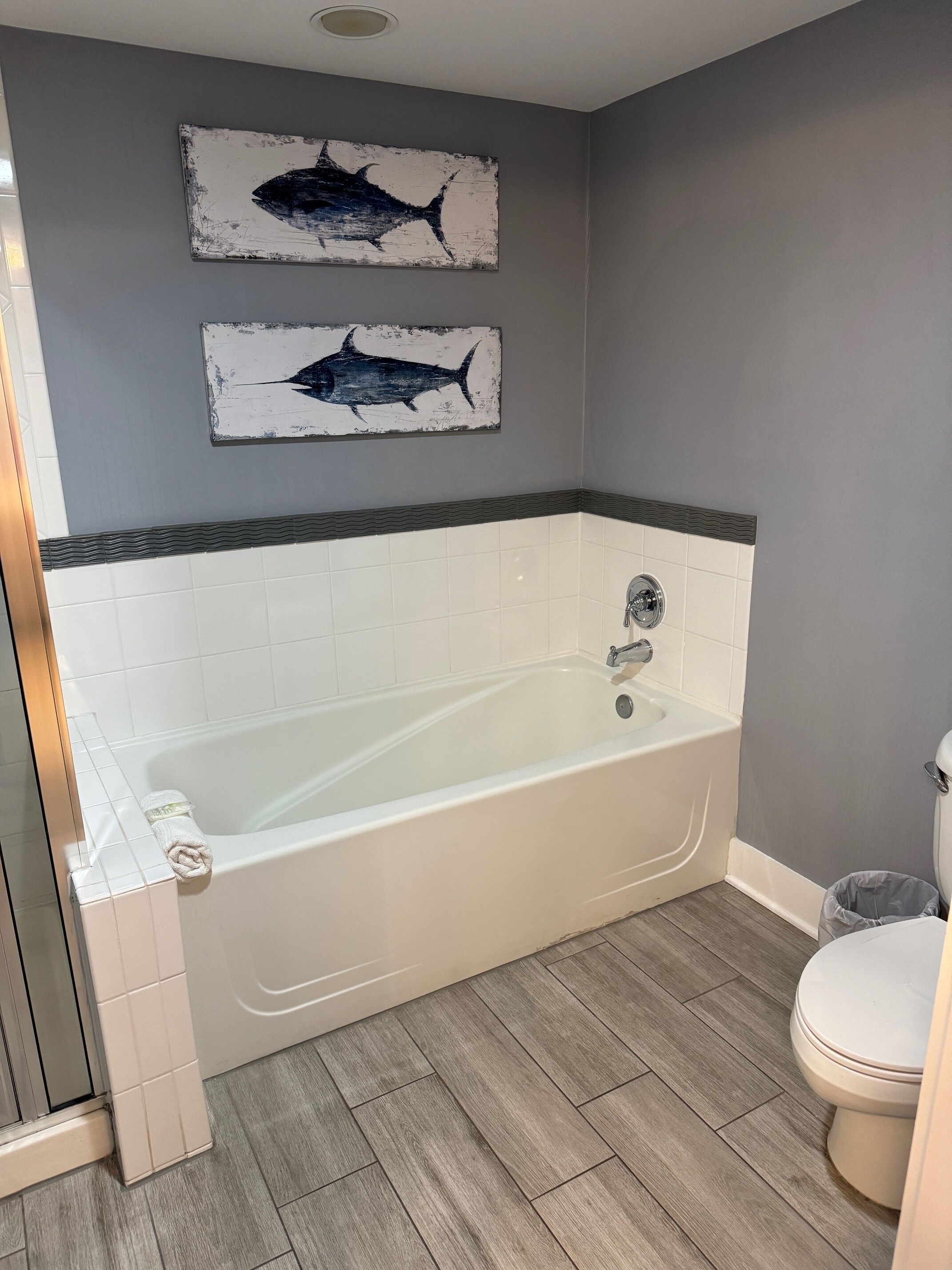 A bathroom with a bathtub, two wall-mounted fish paintings, a toilet, and a shower area.