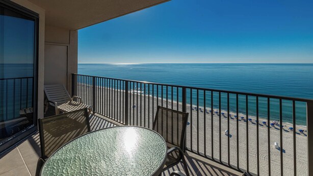 Un balcon avec une table en verre et deux chaises, donnant sur une plage parsemée de parasols et sous un ciel bleu dégagé.