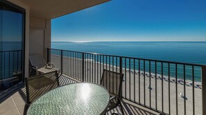 A balcony with a glass table and two chairs, overlooking a beach with umbrellas and a clear blue sky. - Sip, Sun, & Sea Gulf-Front Stay 2 Bedroom Phoenix X 1415 (Orange Beach)