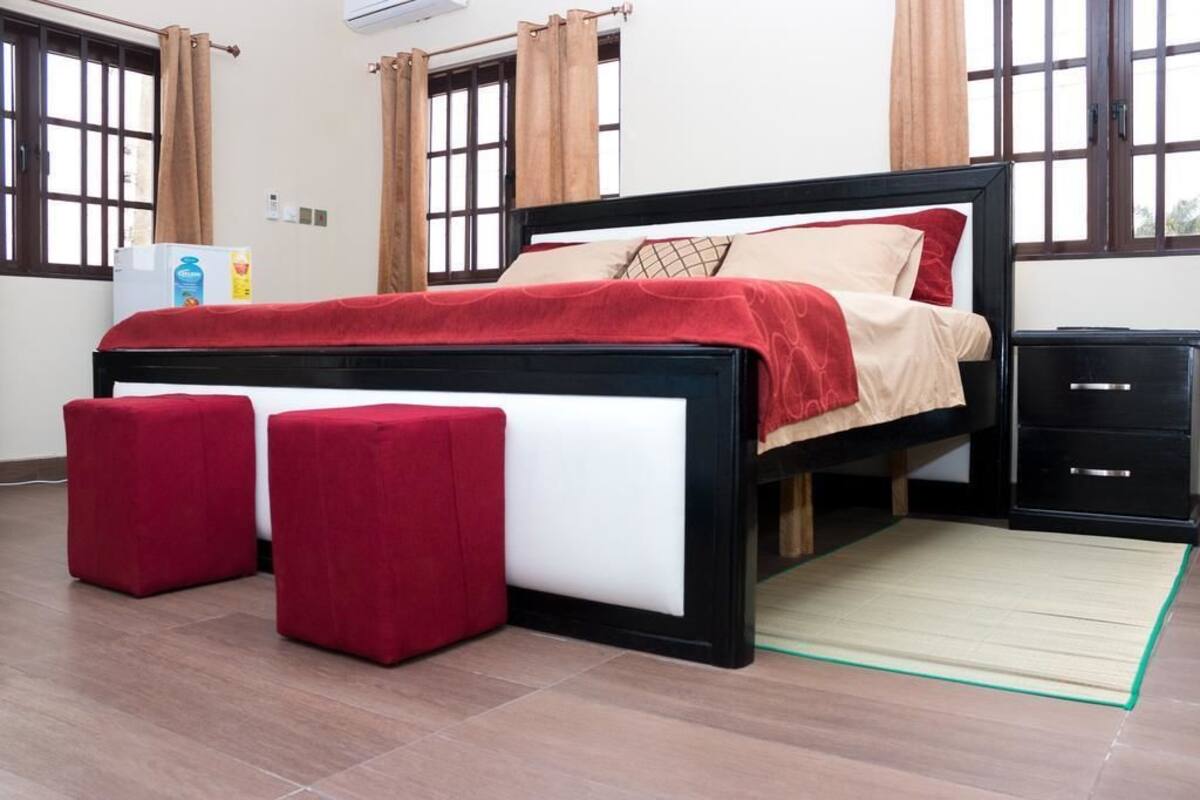 Een slaapkamer met een bed, twee rode poufs, een nachtkastje en een klein kastje.