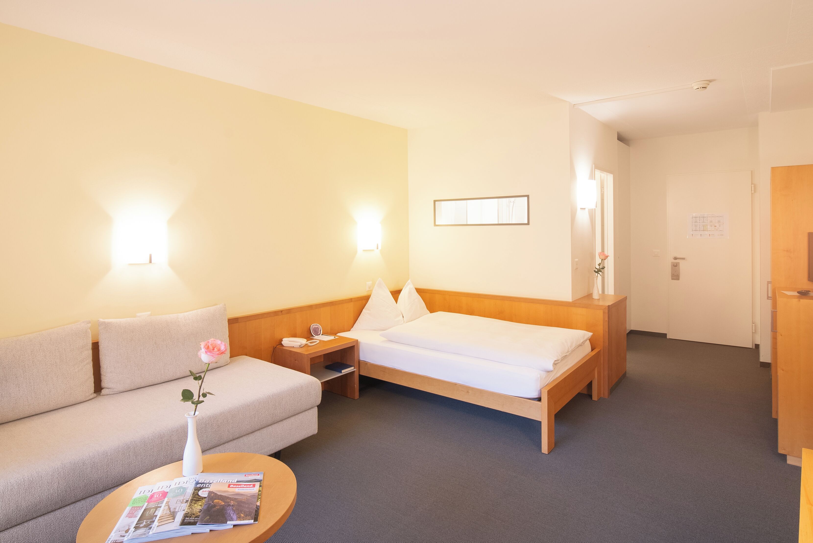 Una habitación de hotel con una cama, un sofá, una mesita de noche con lámpara y una pequeña mesa con un jarrón y revistas.