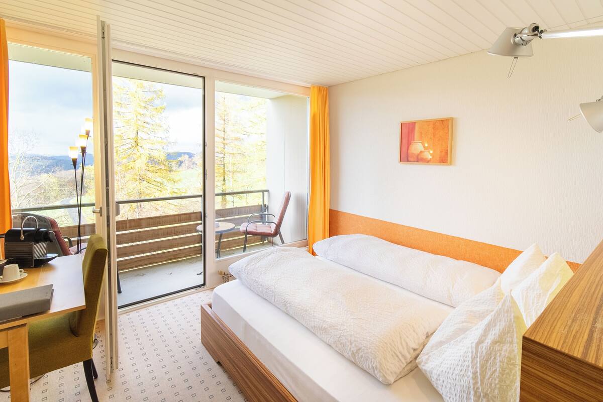A hotel room with a bed, a desk, a chair, and a balcony with a view of trees.