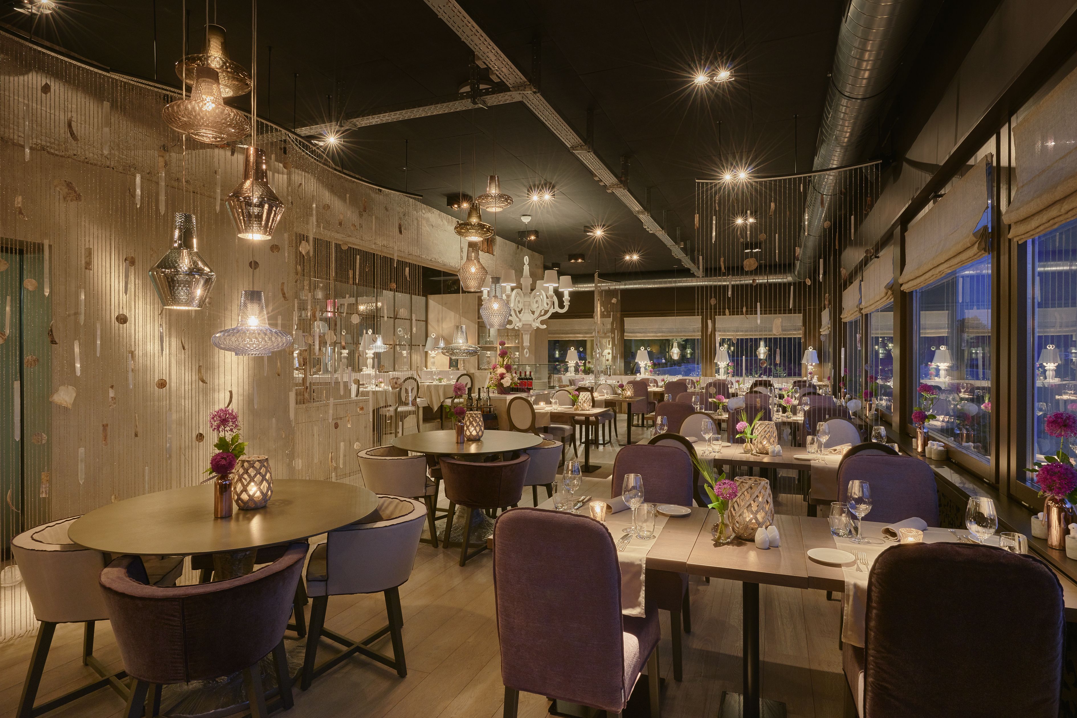 Un restaurant avec des tables rondes, des chaises violettes, des luminaires suspendus et une vue dégagée grâce à de grandes fenêtres.