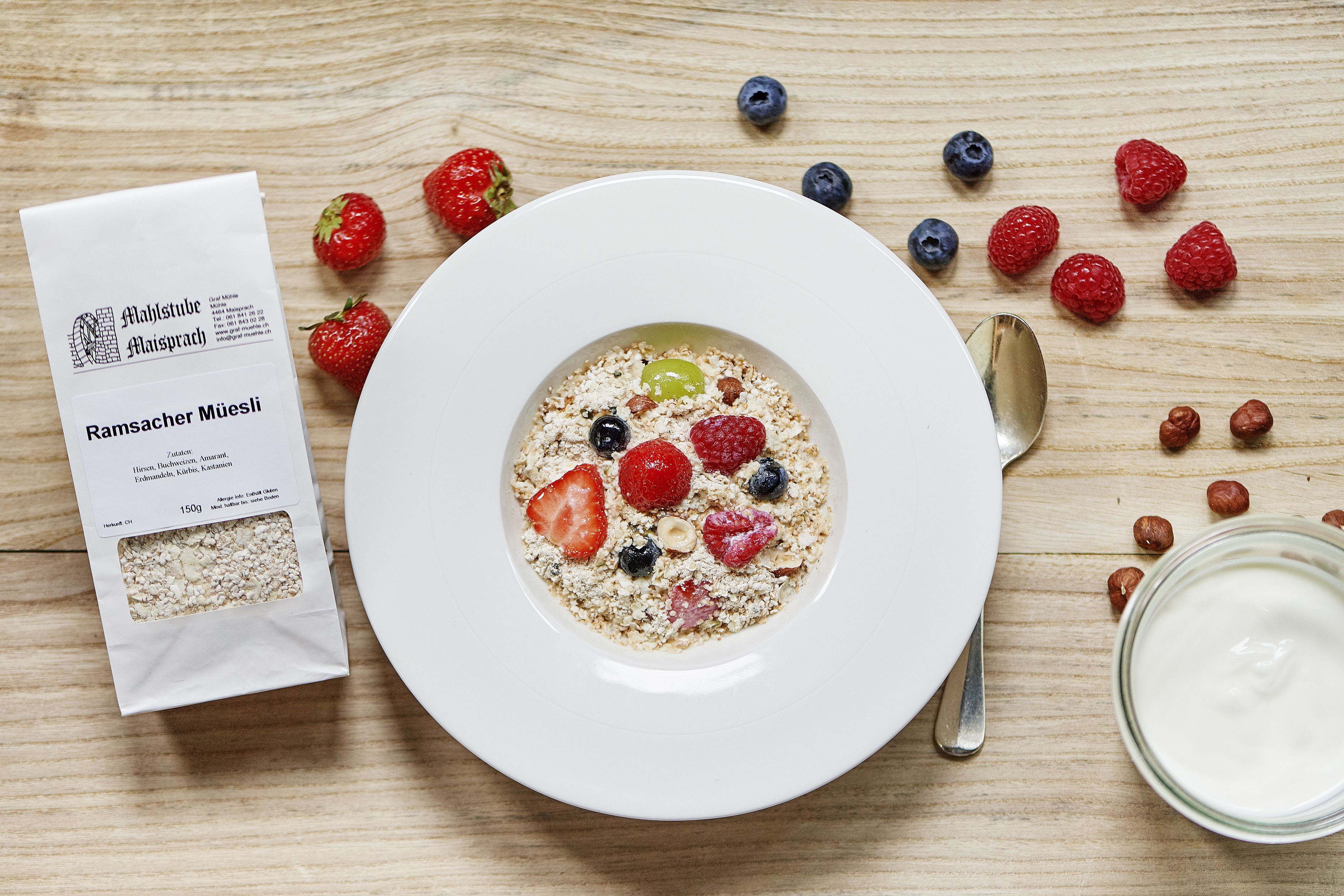 Eine Schüssel mit cerealia, Erdbeeren, Blaubeeren und Himbeeren, ein Löffel, ein Glas Milch und eine Getriebepackung auf einem Holztisch.