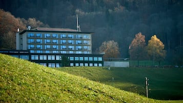 Un edificio de varios pisos con techo plano y numerosas ventanas, ubicado en una colina cubierta de césped con árboles al fondo.