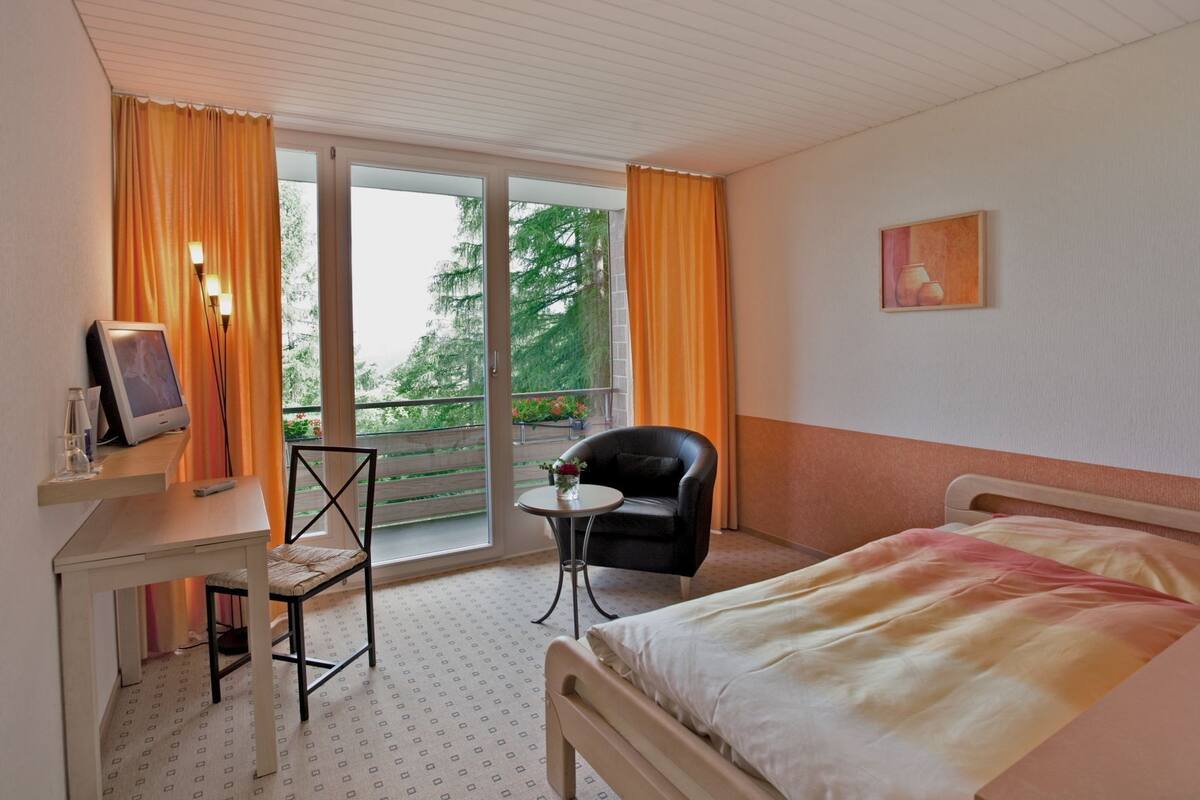 A hotel room with a bed, a desk with a computer, a chair, and a balcony with orange curtains.