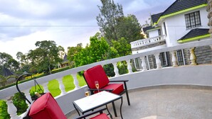 A balcony with red chairs and a table, overlooking a garden and a building with a white facade and black roof. - Sosa Cottages (Gisambai)